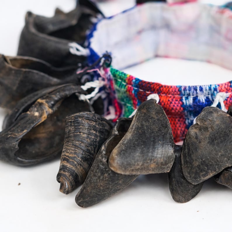 Puede incluir: Primer plano de un collar con cuentas oscuras en forma de cuerno. Las cuentas est&aacute;n ensartadas con una banda de tela estampada de colores rojo, azul, verde y blanco. El collar est&aacute; sobre un fondo blanco.