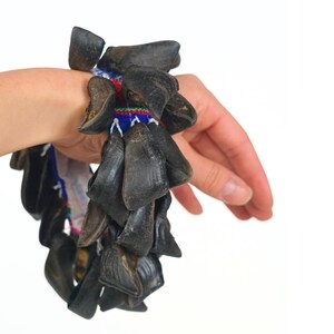 Puede incluir: Una pulsera con m&uacute;ltiples cuentas oscuras en forma de pezu&ntilde;a. Las cuentas est&aacute;n unidas a una colorida banda tejida con detalles en rojo, azul y blanco. La pulsera se muestra en una mu&ntilde;eca sobre un fondo blanco.