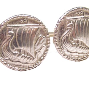 Può includere: Un paio di gemelli d'argento con un design dettagliato di una nave vichinga con una vela. I gemelli sono rotondi e hanno un bordo perlato.
