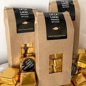 May include: Three brown paper bags with a window showing gold foil wrapped chocolates inside. The bags have black labels with the text "LA LA LAND ORGANICS RAW CACAO" and "Crafted in Western North Carolina". One bag has a label with the text "LA LA LAND ORGANICS .COM".