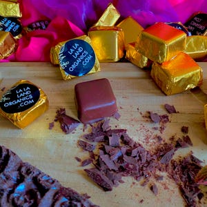 May include: A wooden tray displays chocolate squares, some wrapped in gold foil. One unwrapped square sits next to chocolate shavings. Several wrapped squares have a black and white sticker that reads "LA LA LAND ORGANICS .COM". A pink and purple fabric is in the background.