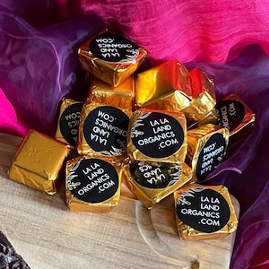 May include: A pile of small, square, gold-wrapped chocolates with black labels that say "LA LA LAND ORGANICS .COM".