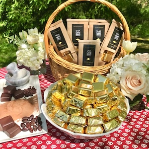 May include: A white wicker basket filled with brown paper bags with the text "La La Land Organic" on them. The basket is sitting on a red and white checkered tablecloth. A white bowl filled with gold foil-wrapped chocolates is in front of the basket.
