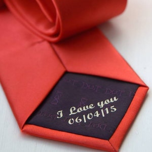 May include: A red silk tie with a dark purple inner lining. The lining has the text "I Love you 06/04/15" embroidered in white.