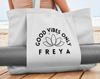 May include: White canvas tote bag with black text that reads "Good Vibes Only Freya" and a lotus flower graphic. The bag is being held by a person and has a gray yoga mat inside.