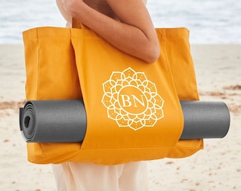 Op de afbeelding: Een gele tote bag met een wit mandala-ontwerp en de letters "BN" in het midden. Een grijze yogamat is in de tas gestopt.