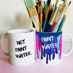 Puede incluir: Dos tazas de cerámica blanca con texto negro. La taza de la izquierda dice "NCT PAINT WATER". La taza de la derecha dice "PAINT WATER" con goteos de pintura de colores. Un montón de pinceles están en un soporte detrás de las tazas.