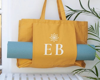 May include: A mustard yellow canvas tote bag with a white starburst design and the initials "EB" printed on the front. The bag has a pocket on the side to hold a teal yoga mat.