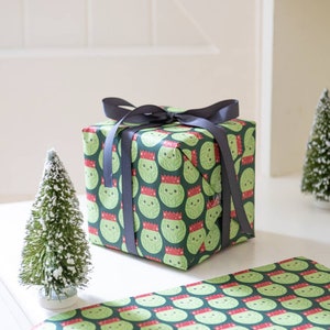 Pode incluir: Um presente embrulhado em papel de embrulho verde com um padr&atilde;o de br&oacute;colis de desenho animado usando chap&eacute;us de Papai Noel. O presente est&aacute; amarrado com uma fita preta. H&aacute; duas pequenas &aacute;rvores de Natal artificiais na mesa.