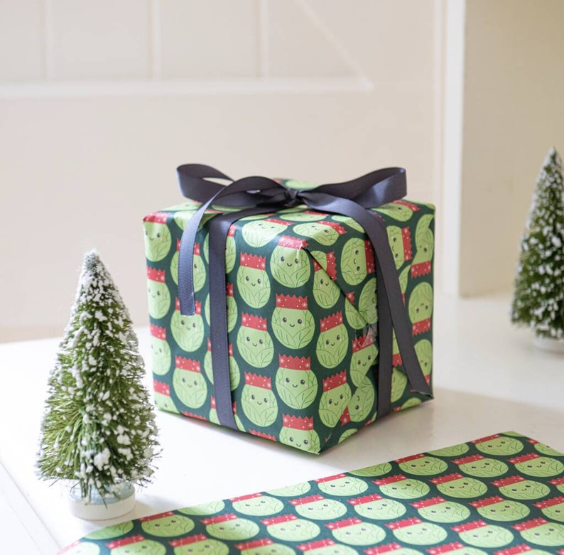 Pode incluir: Um presente embrulhado em papel de embrulho verde com um padr&atilde;o de br&oacute;colis de desenho animado usando chap&eacute;us de Papai Noel. O presente est&aacute; amarrado com uma fita preta. H&aacute; duas pequenas &aacute;rvores de Natal artificiais na mesa.