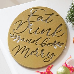 May include: Gold oval-shaped sign with the text "Eat Drink and be Merry" in a decorative font. The sign has a textured surface and is surrounded by a white background.