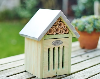 May include: A wooden bee house with a silver metal roof and a light green painted exterior. The house features bamboo tubes for nesting and a decorative metal emblem. The house is sitting on a wooden table.