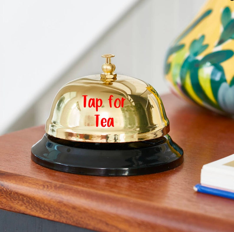 Puede incluir: Una campana dorada brillante con una base negra. El texto rojo en la campana dice "Tap for Tea". La campana est&aacute; sobre una superficie de madera.
