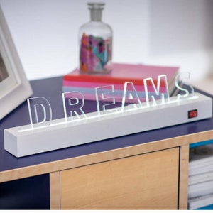May include: White light-up sign with the word "DREAMS" in clear acrylic letters. The sign is on a wooden table with a blue surface.