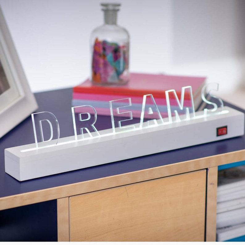 May include: White light-up sign with the word "DREAMS" in clear acrylic letters. The sign is on a wooden table with a blue surface.