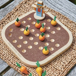 Puede incluir: Un tablero de juego de madera con una superficie de fieltro marrón y 49 agujeros. El tablero tiene un borde marrón claro y el texto "Happy Birthday James" está impreso en el tablero. Hay 10 pequeñas zanahorias amarillas y naranjas con tapas verdes en los agujeros. Una pequeña figura de espantapájaros de madera está en la parte superior del tablero.