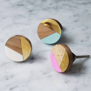 May include: Three round wooden cabinet knobs with geometric designs. The knobs are painted in different colors: white, light blue, and pink. Each knob has a gold accent.