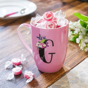 Puede incluir: Una taza de cerámica rosa llena de caramelos envueltos. La taza presenta un diseño floral y una gran letra negra "G". La taza está sobre una superficie de madera, con un plato y una cuchara en el fondo.
