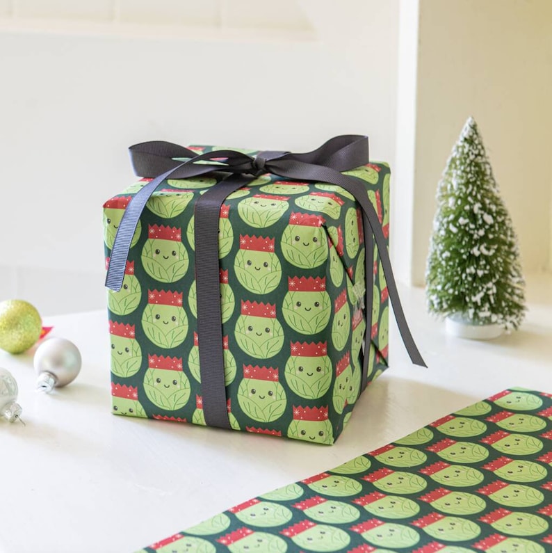 Pode incluir: Um papel de embrulho verde e vermelho com um padr&atilde;o de br&oacute;colis de desenho animado usando chap&eacute;us de Papai Noel. O presente &eacute; embrulhado com uma fita preta.
