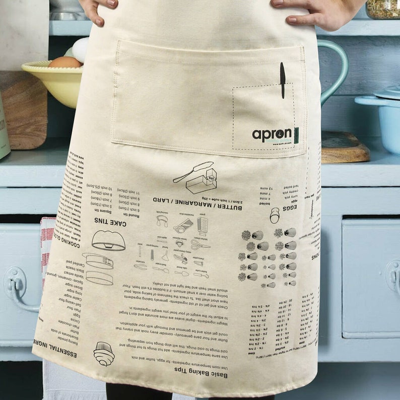 Puede incluir: Un delantal beige con un bolsillo grande y una impresi&oacute;n negra de consejos y medidas para hornear. El delantal tiene la palabra "apron" impresa en el bolsillo.