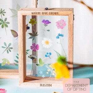 以下が含まれることがあります： 押し花と葉が入った木製フレーム。上部に「WHERE LOVE GROWS」、下部に「31.05.1994」の文字。フレームには、様々な色の花と葉が収められています。