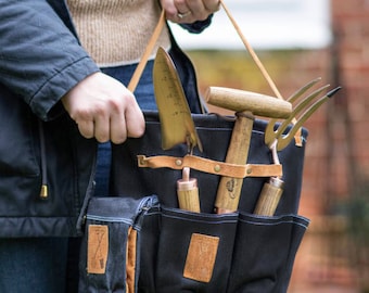 May include: A black canvas gardening tool bag with multiple pockets and a leather strap. The bag is filled with gardening tools, including a trowel, a hand rake, a pair of shears, and a measuring tool.