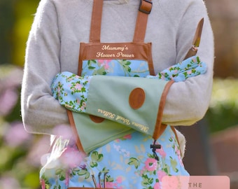 May include: A gardening apron, gloves, and tool set. The apron is blue with a floral pattern and brown trim. The gloves are light blue with a floral pattern. The apron reads "Mummy's Flower Power" and the gloves read "Mums Weed Killers".