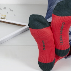 May include: A pair of red and green socks with the text "Cheesy Feet!" on one sock and "Dad's" on the other sock.