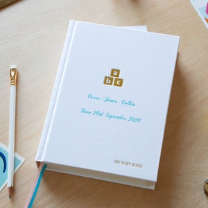May include: A white baby book with a gold and blue design. The book has the text "My Baby Book" on the bottom and "Oscar James Collins Born 20th September 2020" on the front cover.