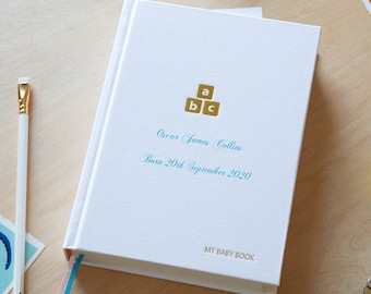 May include: White baby book with a gold and blue design. The book has the text "My Baby Book" on the bottom and "Oscar James Collins Born 20th September 2020" on the front cover.