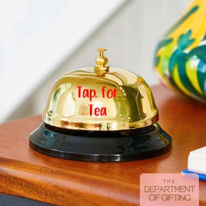 Puede incluir: Una campana dorada brillante con una base negra. La campana tiene la inscripci&oacute;n "Tap for Tea" en letras rojas. Est&aacute; colocada sobre una superficie de madera. El logotipo "The Department of Gifting" est&aacute; en la esquina inferior derecha.
