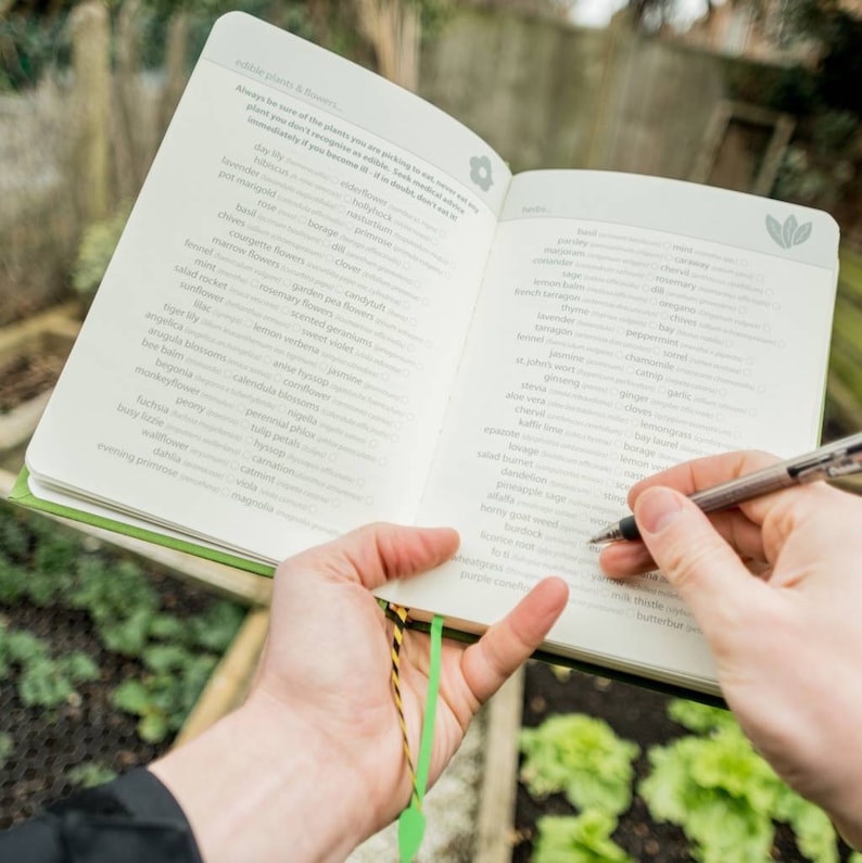 May include: An open book with a green cover and a bookmark. The book lists edible plants and flowers, including day lily, elderflower, lavender, and pot marigold. The book also lists edible herbs, including rosemary, garlic, chives, and lemongrass.