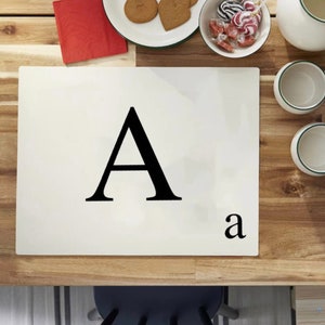 May include: A cream-coloured rectangular tile featuring a large black letter "A" and a smaller "a" in the bottom right corner. The tile is on a wooden table, with biscuits, sweets, and two mugs.