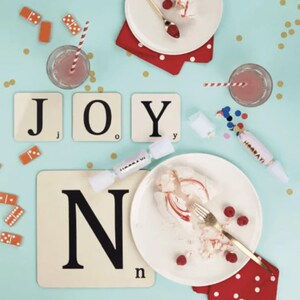 May include: Four white square coasters with black letters spelling out "JOY" and a fifth coaster with the letter "N". The coasters are on a light blue surface with confetti and dominoes. There is a white plate with a fork and a dessert on it.
