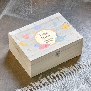 May include: A wooden keepsake box with a light wood finish and a colourful design featuring clouds, stars, and moons. The box has a hinged lid and a silver clasp. The text on the box reads "Felix Lewis Dream Big Little One".