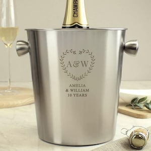 Può includere: Secchiello per champagne in argento con un'iscrizione personalizzata: "A&W Amelia & William 10 Years". L'iscrizione è circondata da una ghirlanda a forma di cuore.
