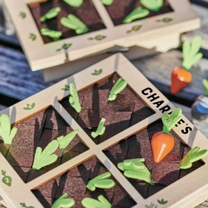 May include: A wooden game board with the text "Charlie's" printed on it. The board has four squares, each with a brown felt-like surface and green felt-like leaves. There are two orange felt-like carrots on the board.