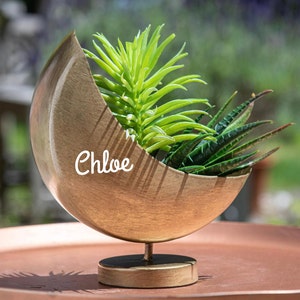 May include: A gold metal planter shaped like a crescent moon with a stand. The planter is filled with green succulents and the name "Chloe" is written on the side.