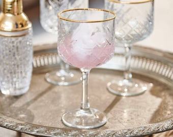 May include: A clear glass cocktail shaker with a gold-colored lid and three stemmed glasses with gold rims. One glass contains a pink drink and ice. The glasses and shaker sit on a silver-colored tray.
