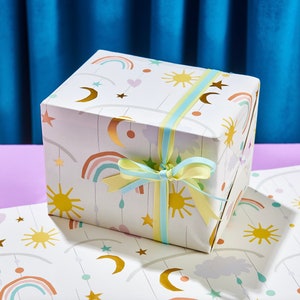 K&ouml;nnte beinhalten: Wei&szlig;e Geschenkpapier mit einem bunten Design aus Regenb&ouml;gen, Sternen, Monden und Sonnen. Das Papier ist mit einem blauen und gelben Band gebunden.