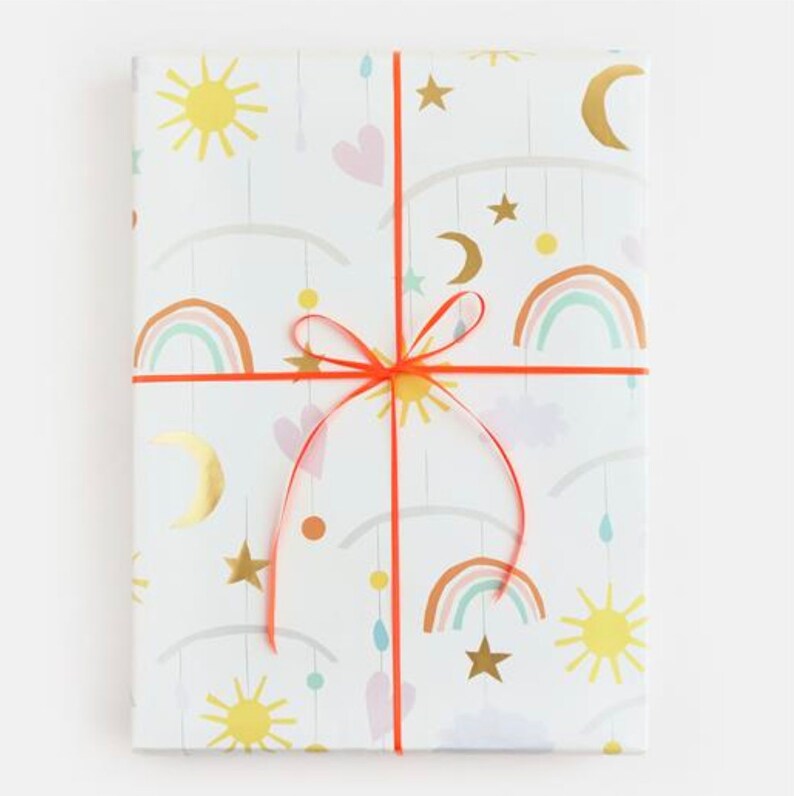 K&ouml;nnte beinhalten: Wei&szlig;e Geschenkpapier mit einem sich wiederholenden Muster aus Regenb&ouml;gen, Sternen, Monden und Sonnen. Das Papier ist mit einem roten Band gebunden.