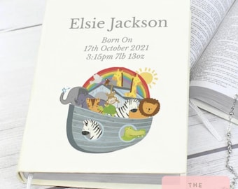 Op de afbeelding: Crèmekleurig baby-aandenkenboek met de naam "Elsie Jackson" en geboortegegevens in grijs gedrukt. De omslag toont een kleurrijke illustratie van dieren op een ark onder een regenboog. Het gewicht van het boek is 3,54 kg.