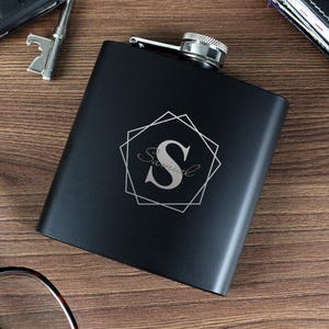 Puede incluir: Una petaca negra con un diseño geométrico plateado y la letra "S" y el nombre "Samuel" grabados en la parte delantera. La petaca tiene una tapa plateada y es de forma rectangular. La petaca está sobre una superficie de madera.