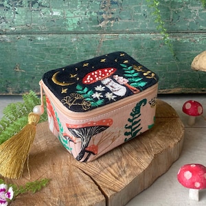 May include: A small, rectangular box with a zipper closure. The box is covered in a pink fabric with an embroidered design of mushrooms, ferns, and stars. The top of the box is black with a gold crescent moon and stars. The box has a gold tassel.