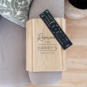 以下が含まれることがあります： 「Reserved for Harry's Cup of Tea」の文字と黒いリモコンが付いた、明るい色の木製ソファアームトレイ。 トレイは長方形で、灰色のソファのアームの上に置かれています。