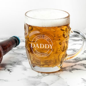 May include: A clear glass beer mug filled with golden beer and white foam. The mug has a handle and the text "DADDY Our Hero!" in a circular design. A brown beer bottle with a blue cap is in the background.