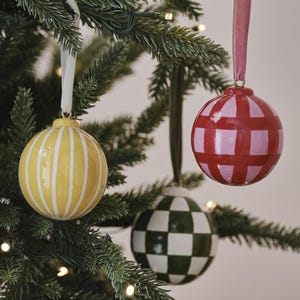 May include: Three decorative Christmas baubles hanging on a green tree. The baubles are round and painted with different patterns: yellow with white stripes, red with pink squares, and green with white checkerboard.
