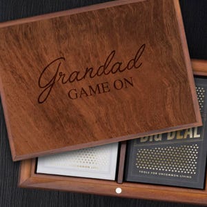 Może przedstawiać: Drewniane pudełko z wygrawerowanym napisem "Grandad GAME ON" na pokrywie. Pudełko jest otwarte, ukazując dwie talie kart. Jedna talia jest biała ze złotymi akcentami, a druga czarna ze złotymi akcentami. Pudełko jest wykonane z ciemnego drewna.
