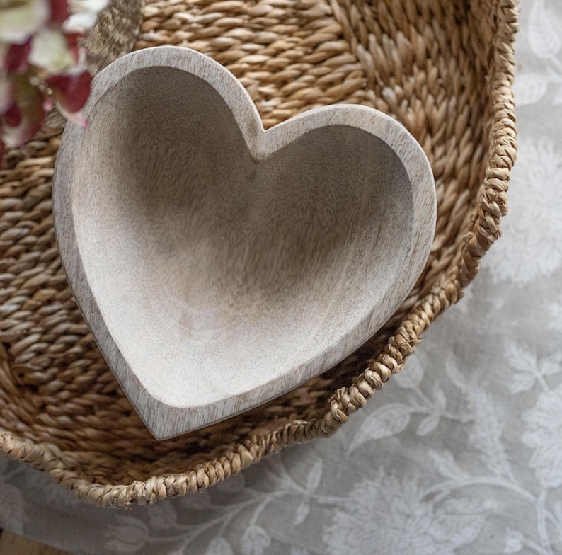 May include: A heart-shaped bowl crafted from light-toned wood with a visible grain pattern. The bowl features a smooth, hollowed-out interior and a thick, sturdy rim, showcasing the natural texture and pale beige color of the wood material.
