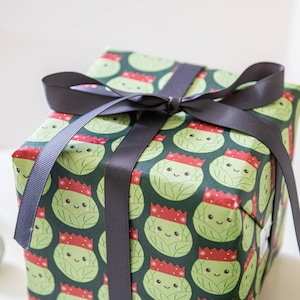 Pode incluir: Uma caixa de presente verde com uma fita preta. A caixa &eacute; embrulhada em papel com um padr&atilde;o de br&oacute;colis de desenho animado usando chap&eacute;us de Papai Noel.
